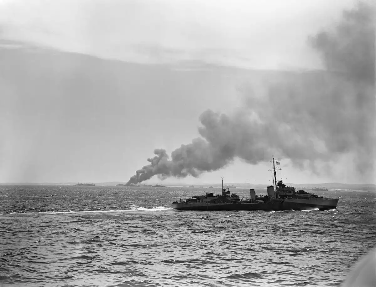 El HMS Eskimo patrulla la zona de desembarco en la costa de Sicilia durante la invasión aliada, en julio de 1943; al fondo, un buque de suministros arde tras ser alcanzado por aviones enemigos.